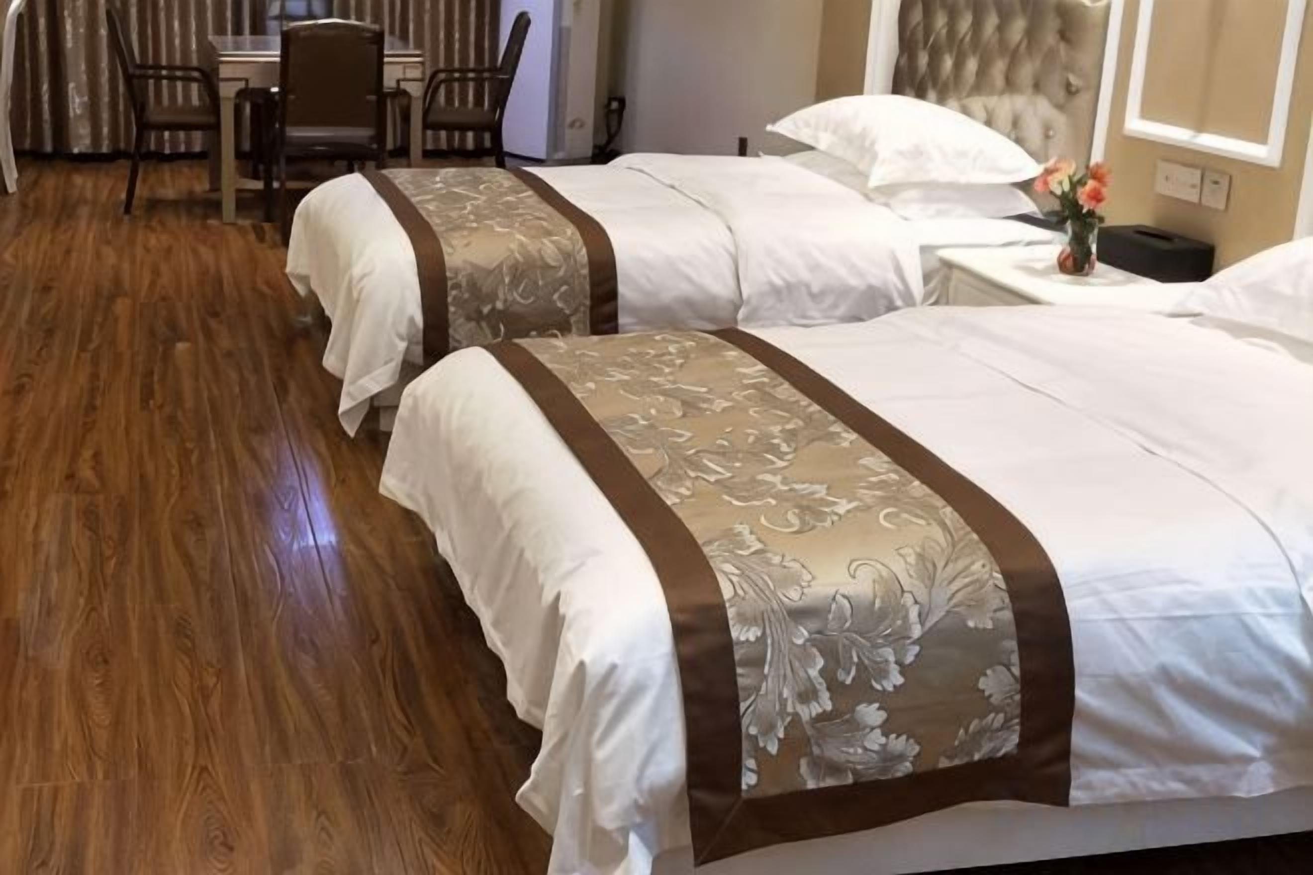 Two beds with matching headboards and footboards, a wooden floor, a small table with chairs, and a nightstand with a vase of flowers.