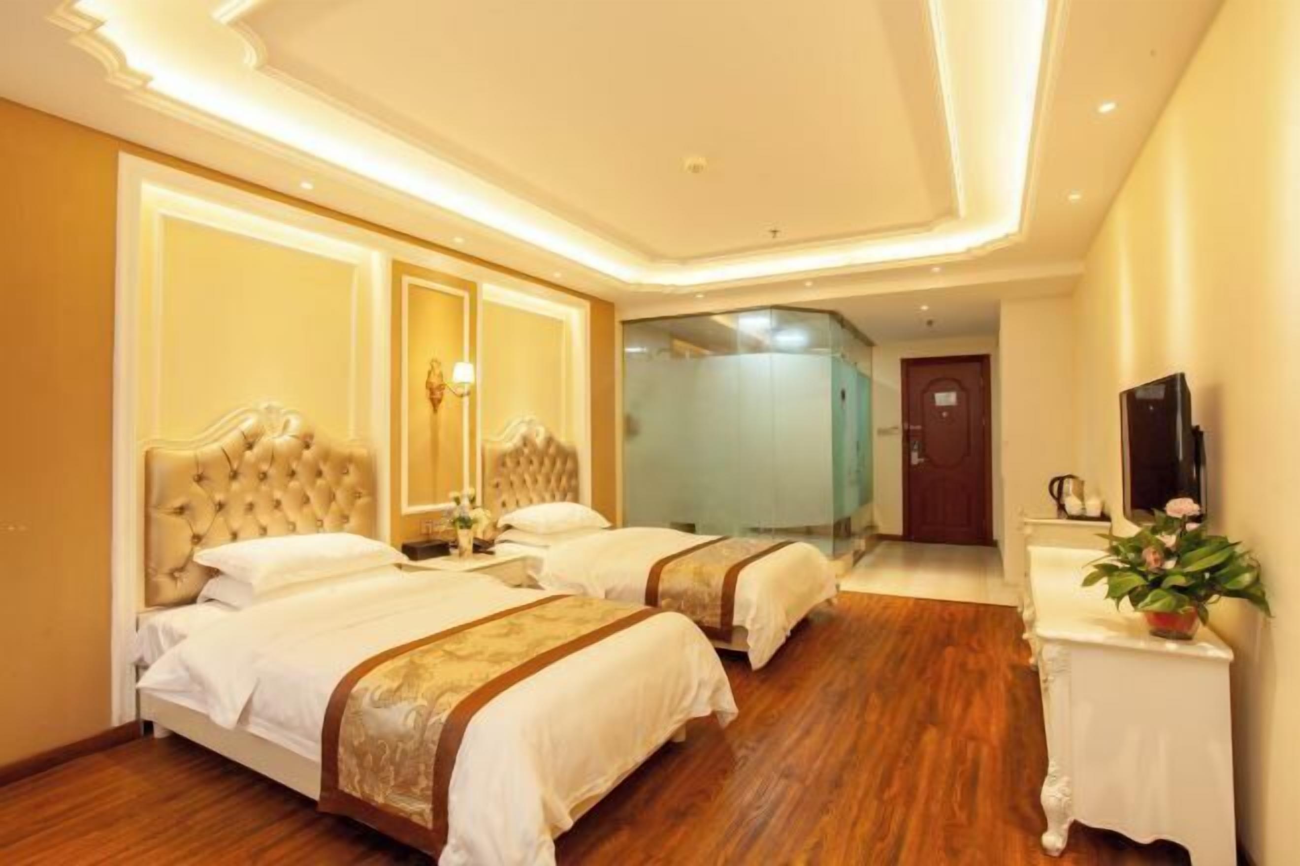 A hotel room with two beds, a wooden floor, a white console table with a plant, and a mirror on the wall.