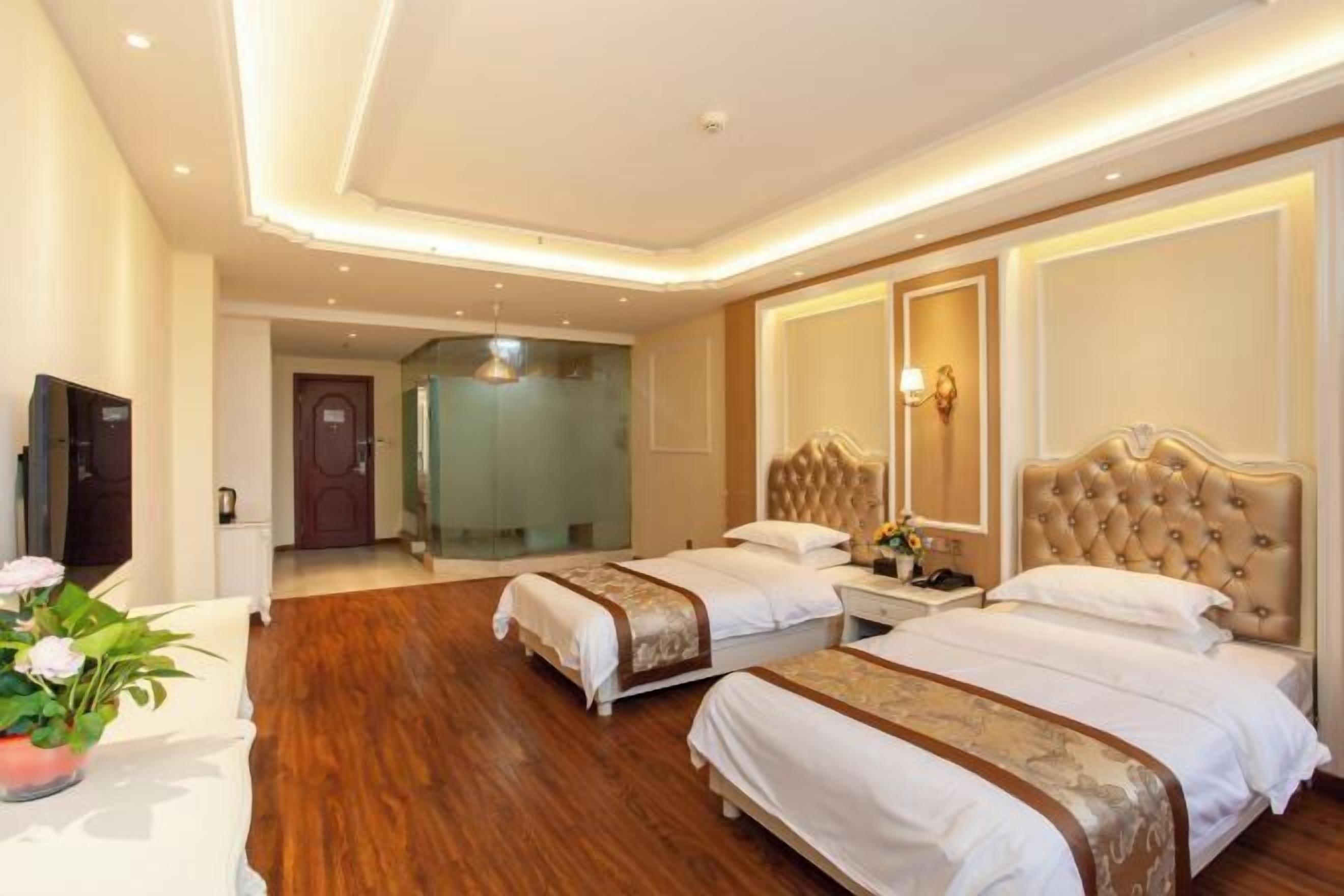 A hotel room with two beds, wooden flooring, a television, and a door leading to a bathroom.