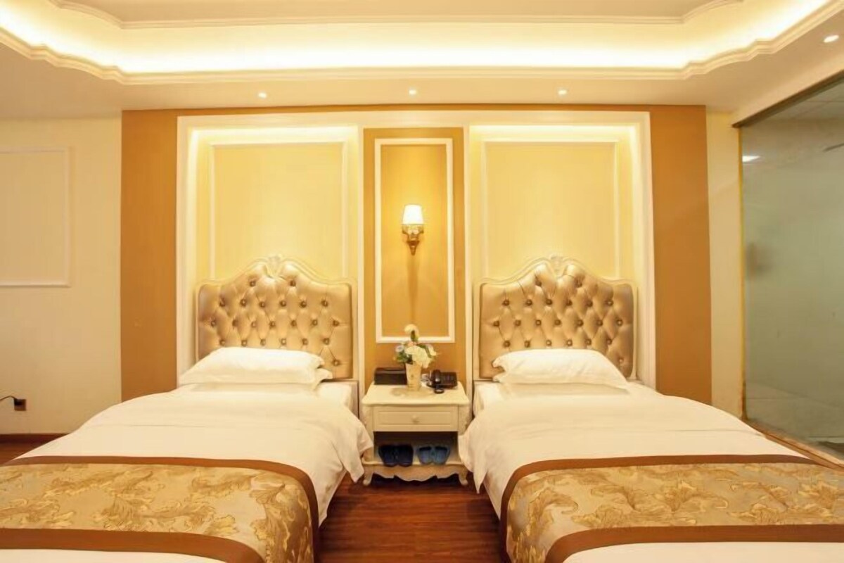A hotel room with two beds, a nightstand, a mirror, and a wall-mounted lamp.