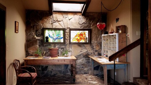 A rustic stone wall with two windows, a wooden desk, a chair, and a staircase.