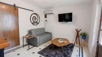 Un espacio habitable compacto con un sofá gris, una mesita de madera, un tapete negro y un televisor montado en la pared.