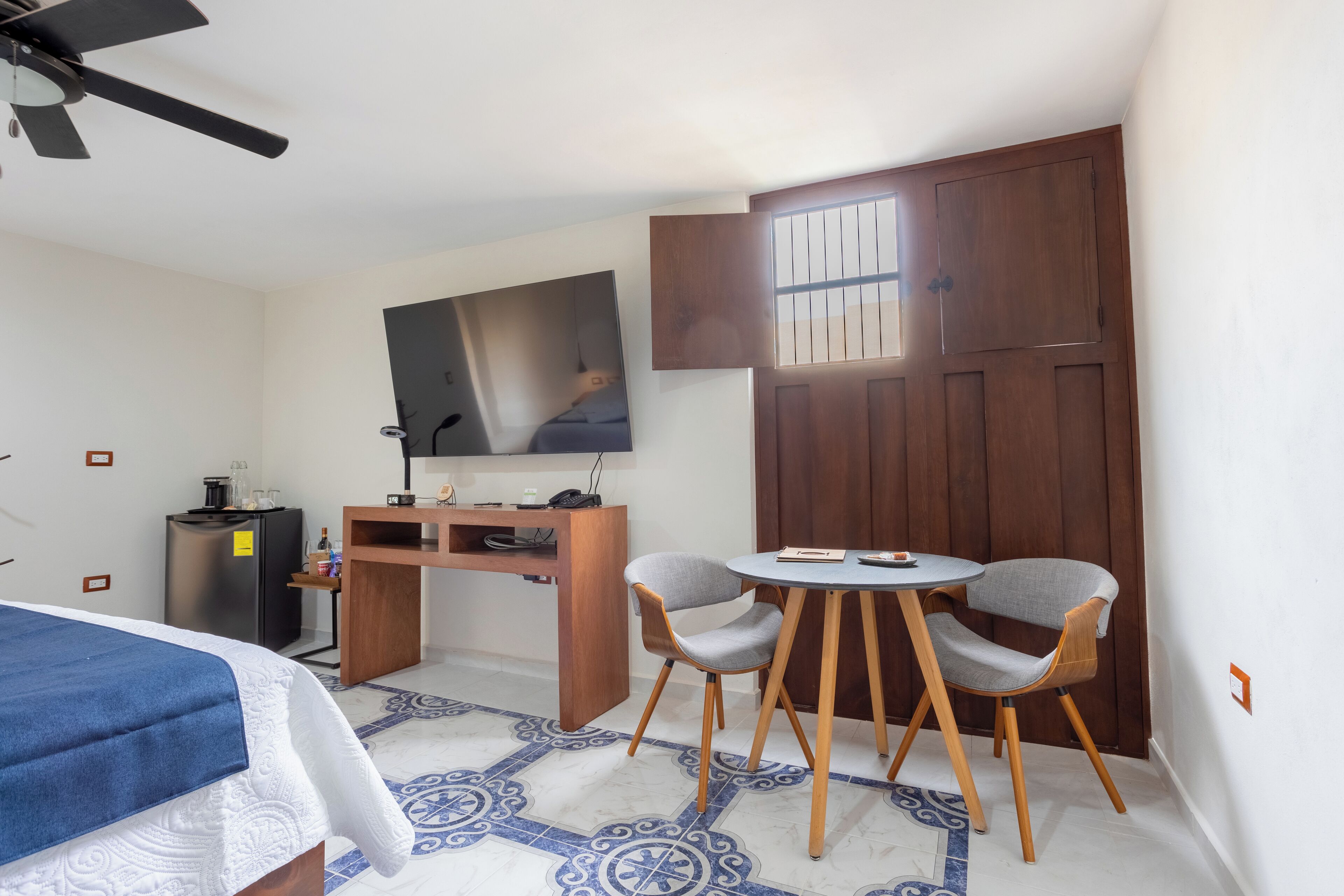 Quarto de hotel com cama, escrivaninha e cadeira, mesinha, frigorífico e janela com grades.