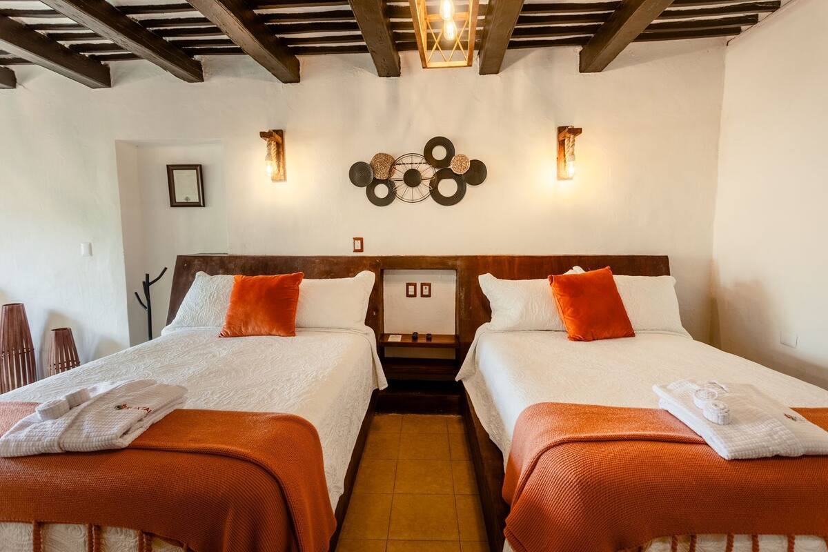 Una habitación de hotel con dos camas, cada una con ropa blanca y almohadas anaranjadas. Hay lámparas de pared y un cabecero de madera.