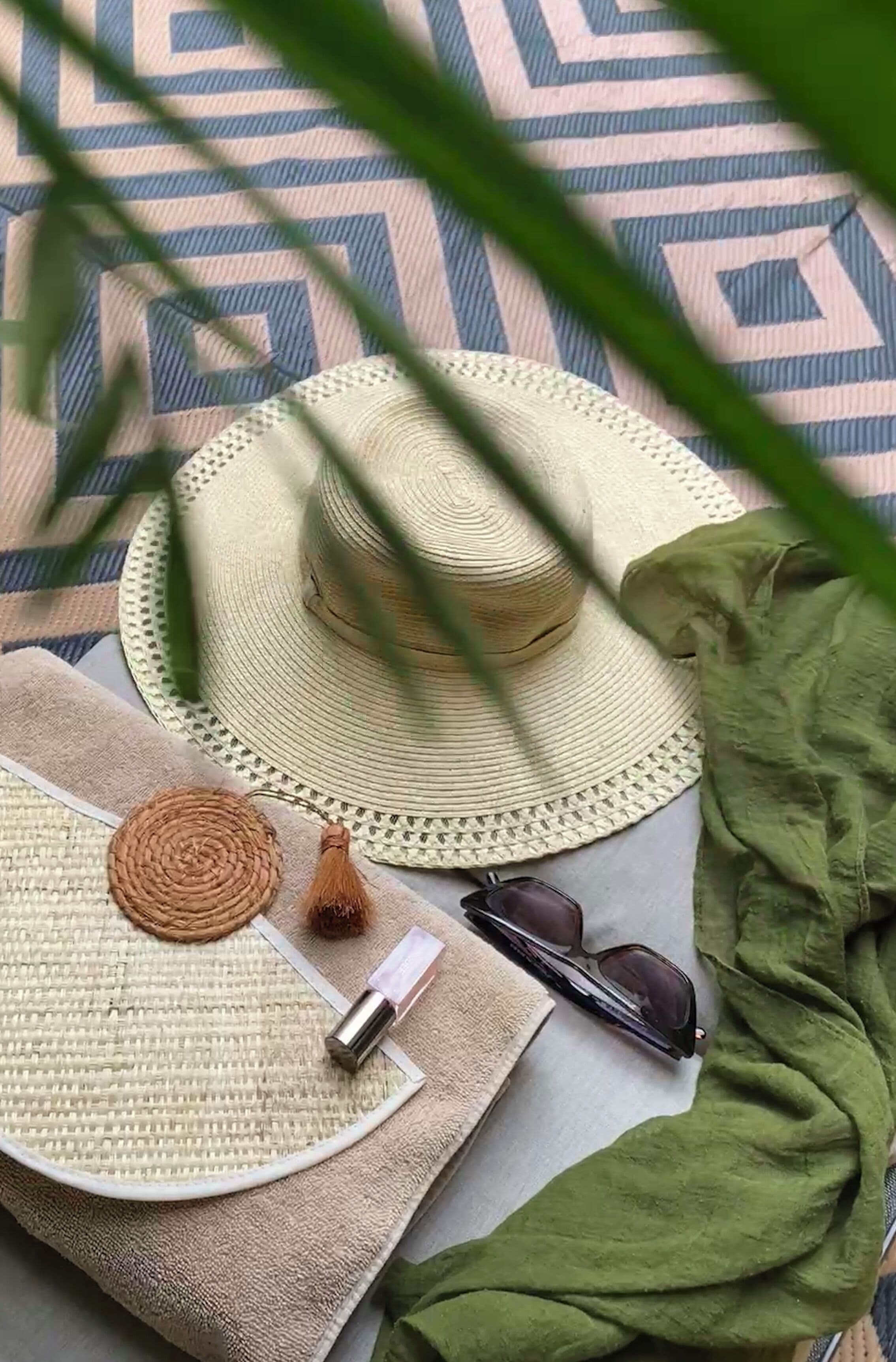 Un chapeau de paille, des lunettes de soleil, un tapis tissé, une petite bouteille et un morceau de tissu vert posés sur une surface à motifs.
