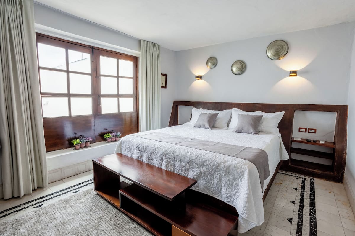 Un dormitorio con una cama grande, un banco de madera, una ventana con cortinas e iluminación en la pared.