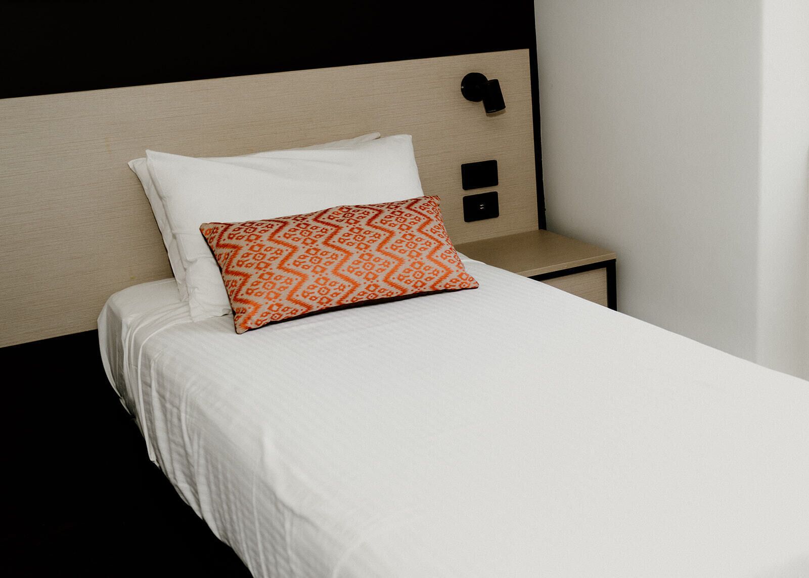 A neatly made bed with white bedding and a patterned orange pillow, a bedside table with a black switch, and a plain wall.