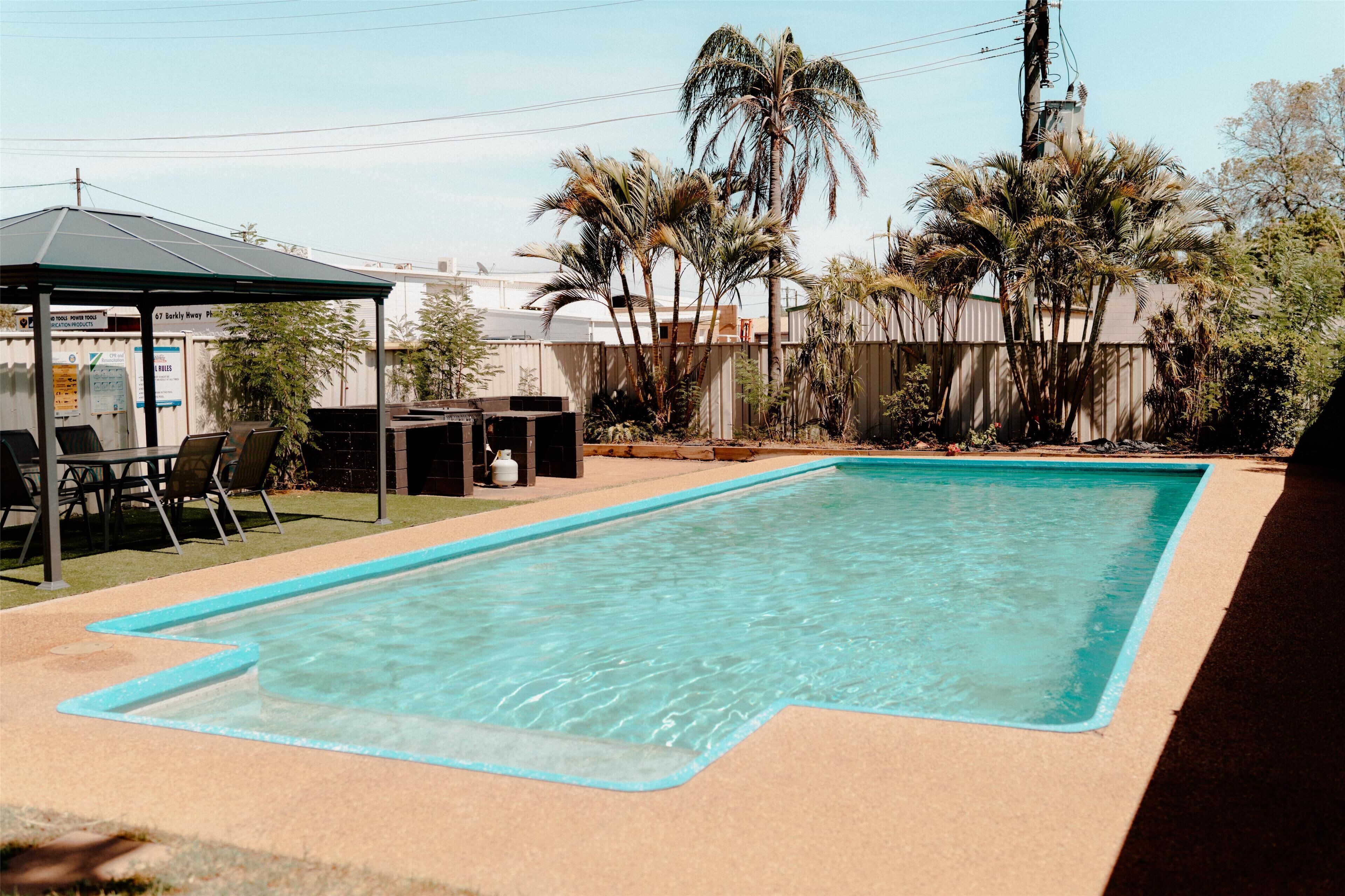 A rectangular swimming pool with a blue edge, surrounded by a concrete deck. There are chairs and a table under a shaded area to the left. Palm trees and a fence are visible in the background.