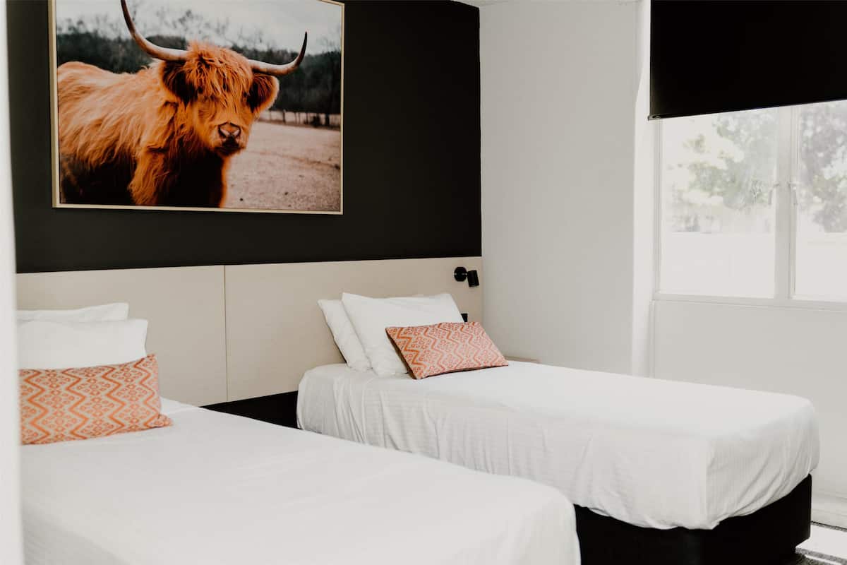 A hotel room with two beds, a large framed picture of a yak, and a window with a view of trees.