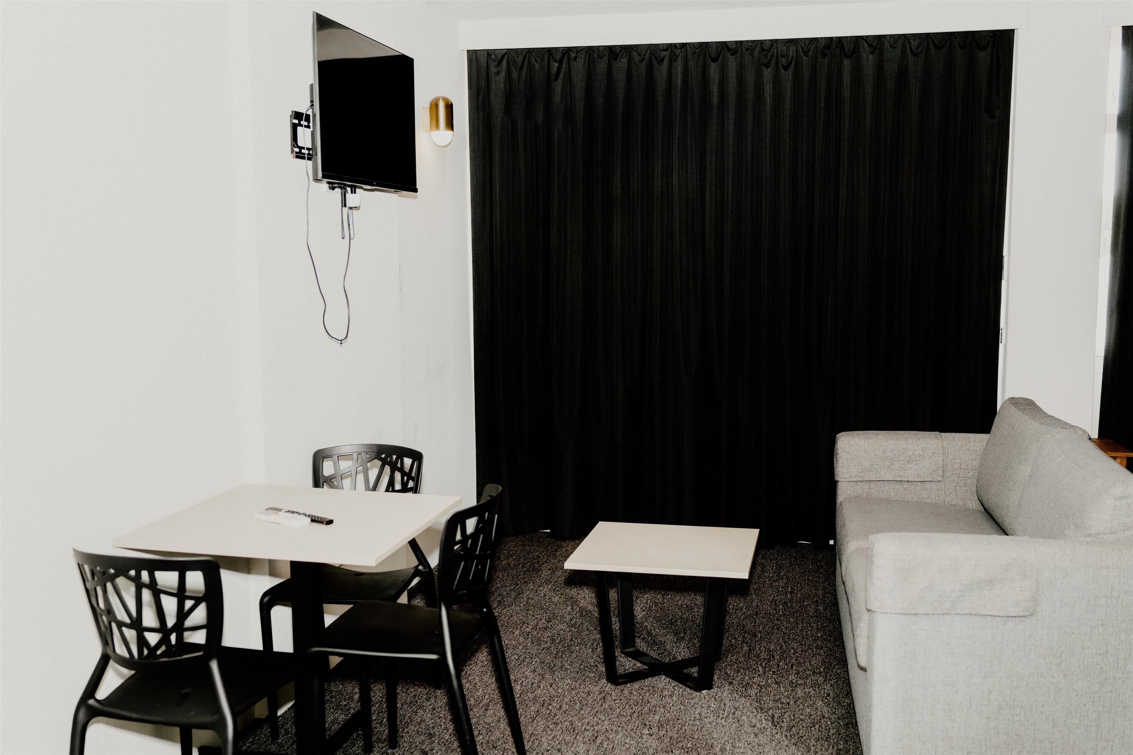 Una habitación pequeña y poco amoblada con una mesa blanca, dos sillas negras, un sofá gris, una cortina negra y un televisor de pared.