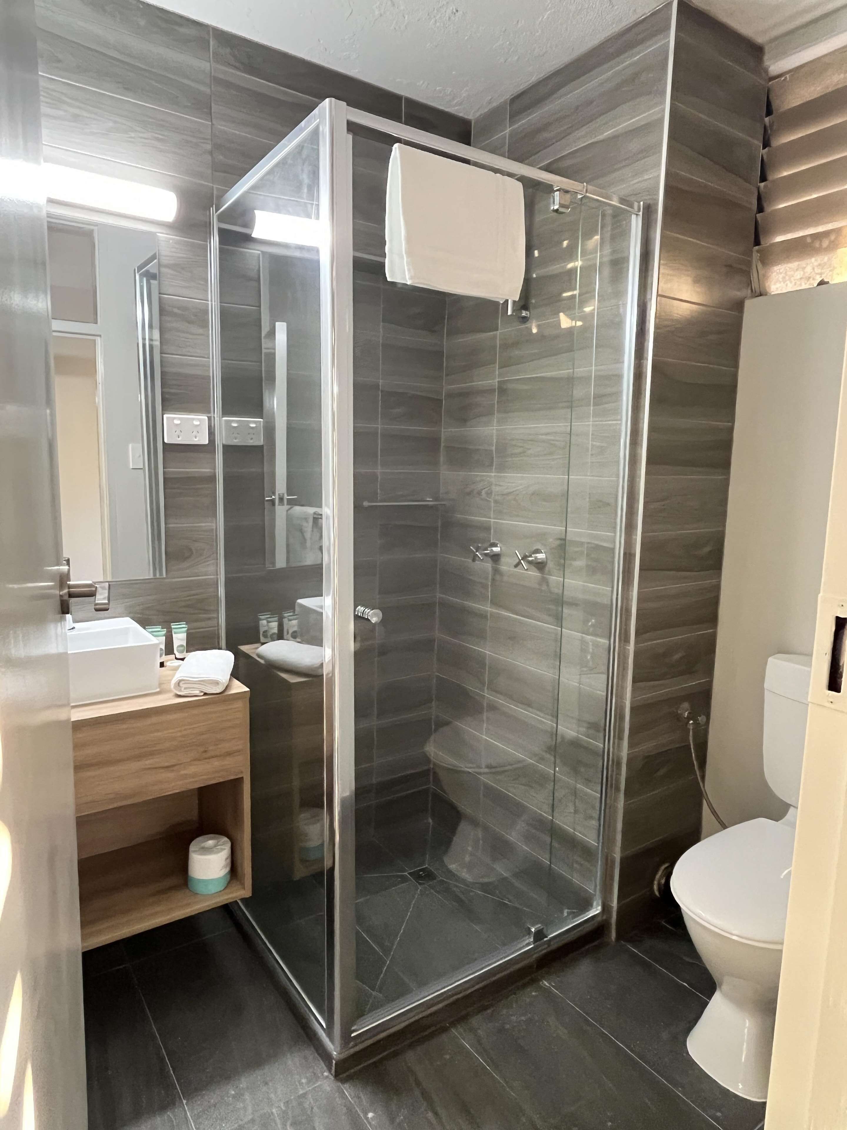 Un baño moderno con ducha de vidrio, tocador de madera, lavabo blanco y un inodoro.