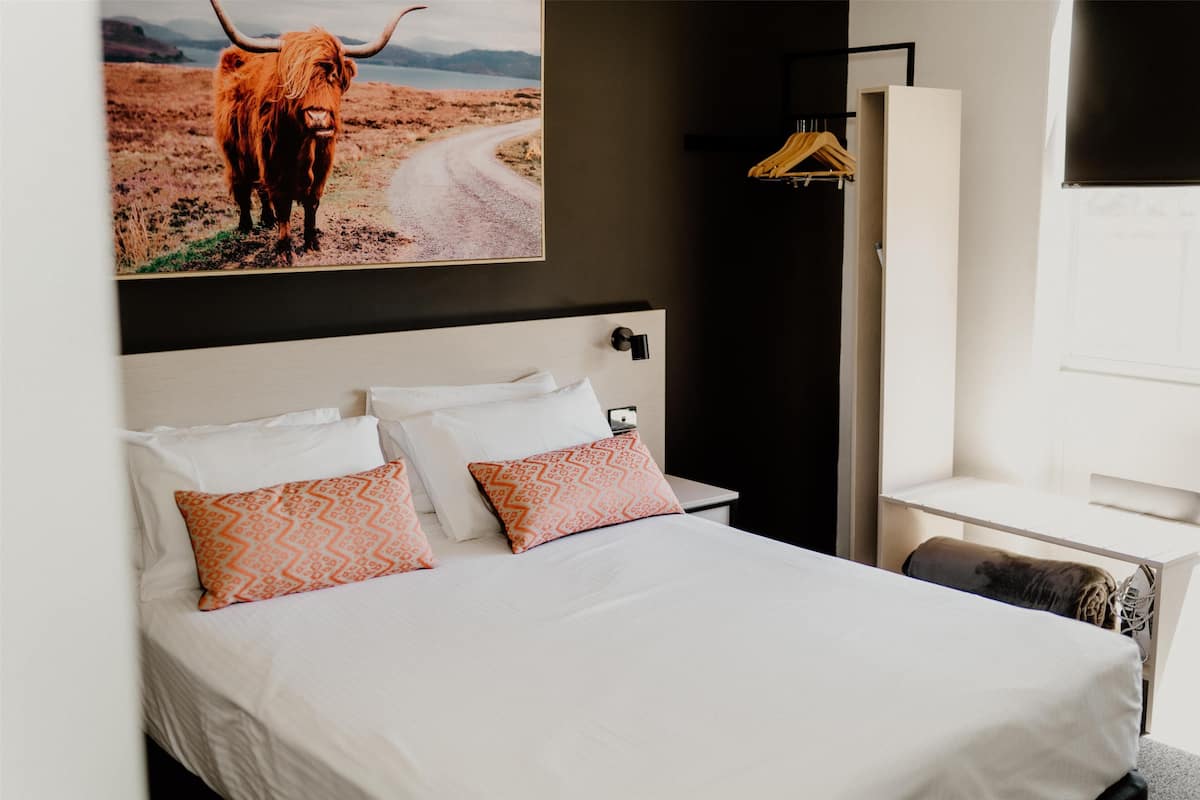 A hotel room with a bed, a nightstand, a wardrobe, and a painting of a cow on a road.