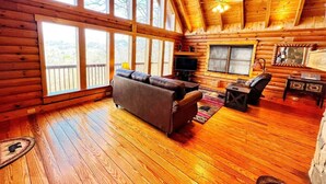 A log cabin interior with a wooden floor, a sofa, a television, and large windows.