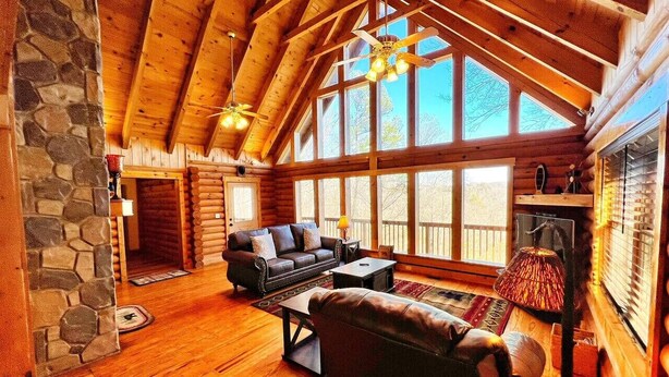 A log cabin interior with a stone wall, wooden floors, a fireplace, a sofa, a coffee table, and large windows.