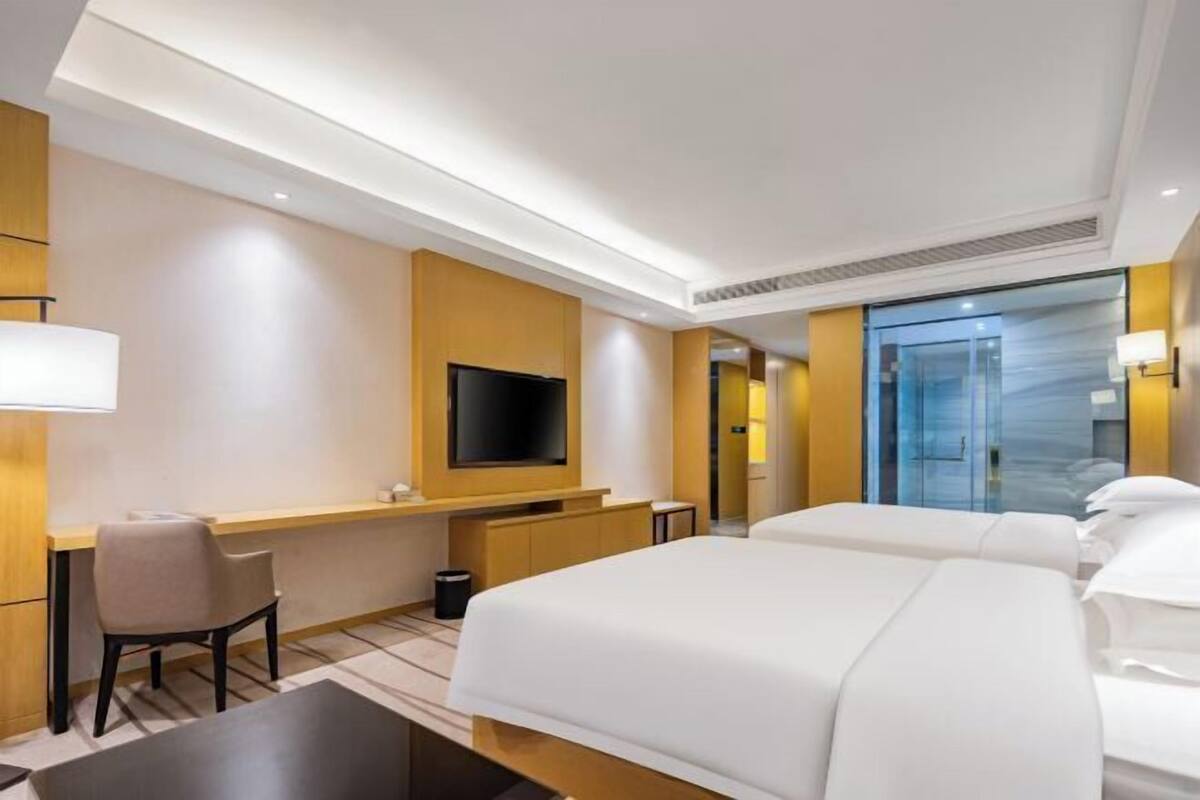 Habitación de hotel con cama, escritorio, silla, televisión y baño visible a través de una puerta de vidrio.