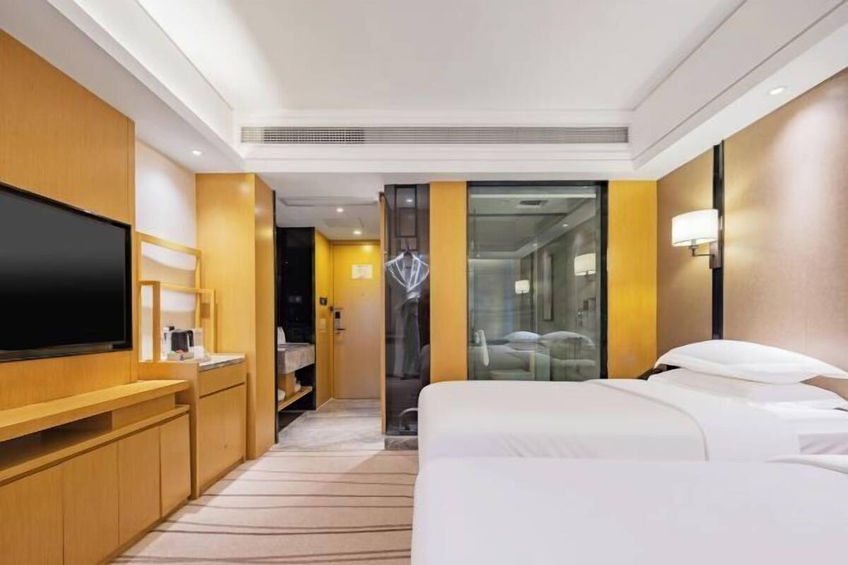 Habitación de hotel con cama, televisor, escritorio, espejo, baño y puerta de vidrio que da a la zona de ducha.