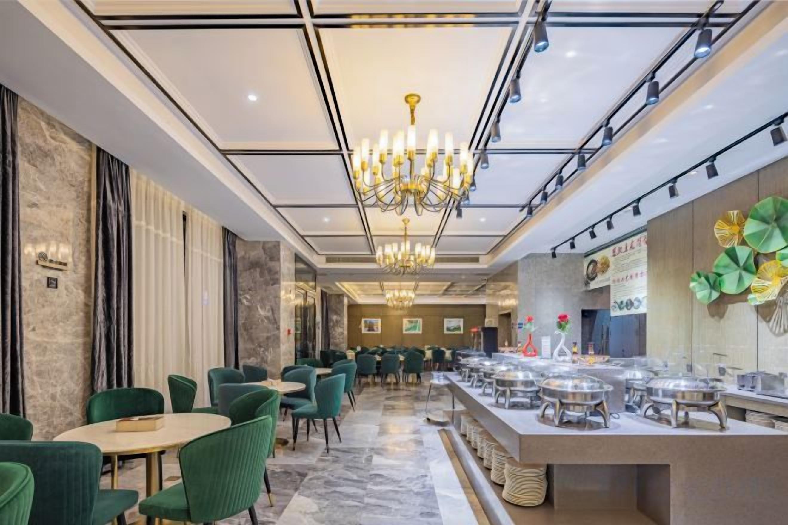 Un comedor de hotel con mesas redondas, sillas verdes, una araña y un mostrador de buffet.