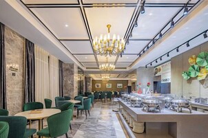 Un comedor de hotel con mesas redondas, sillas verdes, una ara ña y un mostrador de buffet.