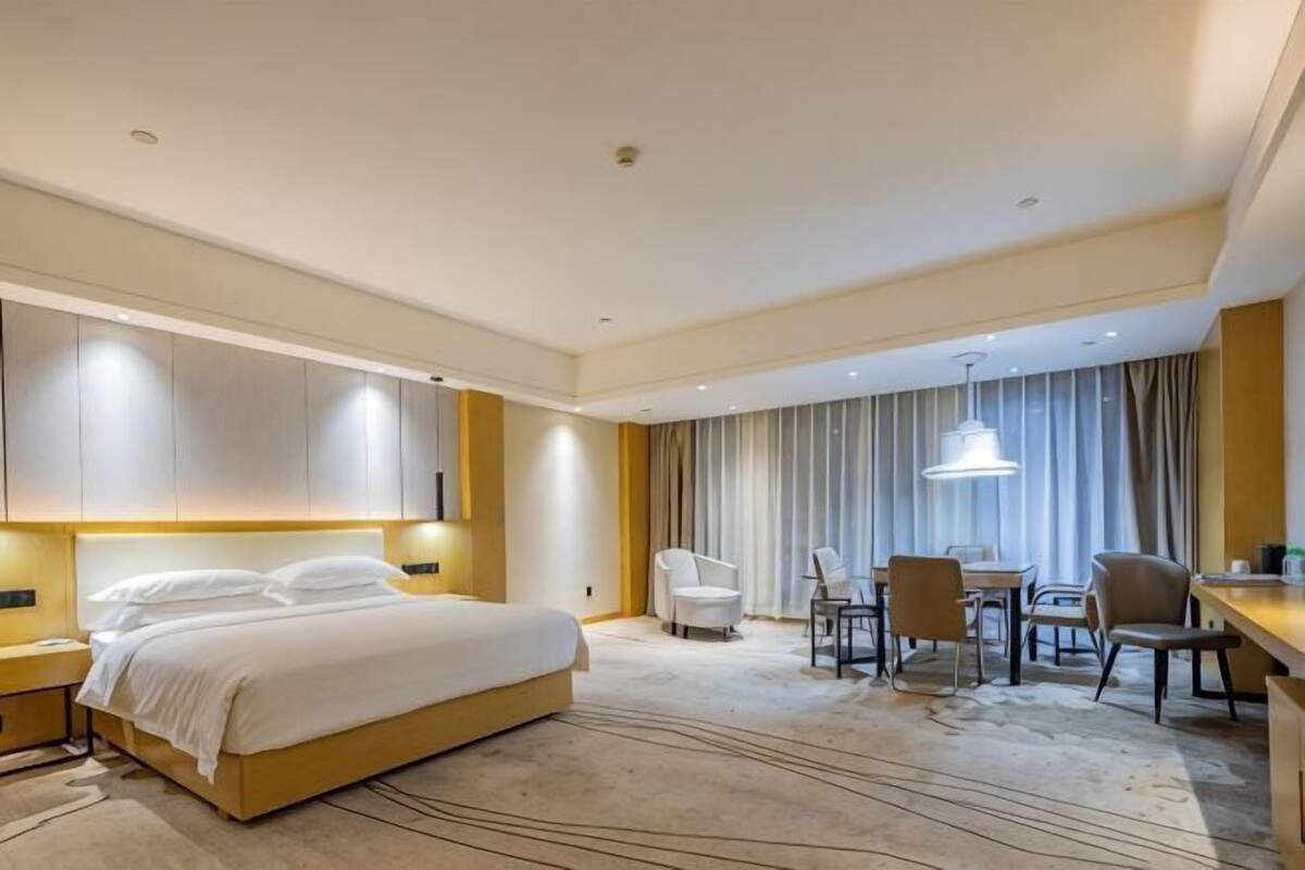 Habitación de hotel con una cama grande, mesitas de noche, un escritorio con sillas, una mesa de comedor y un ventanal con cortinas.