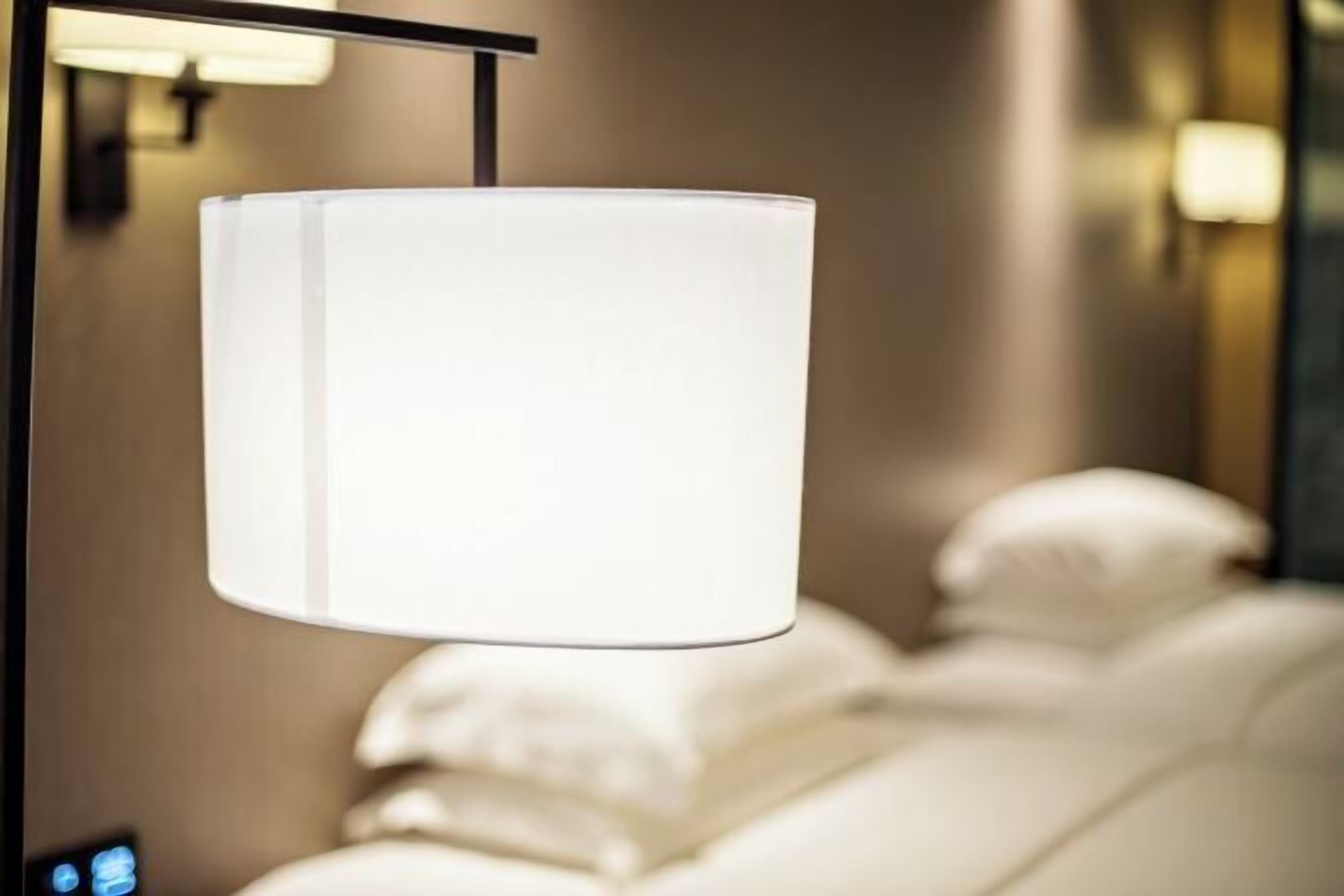 Una habitación con una lámpara colgante cilíndrica, una cama con ropa blanca y un panel de control instalado en la pared.