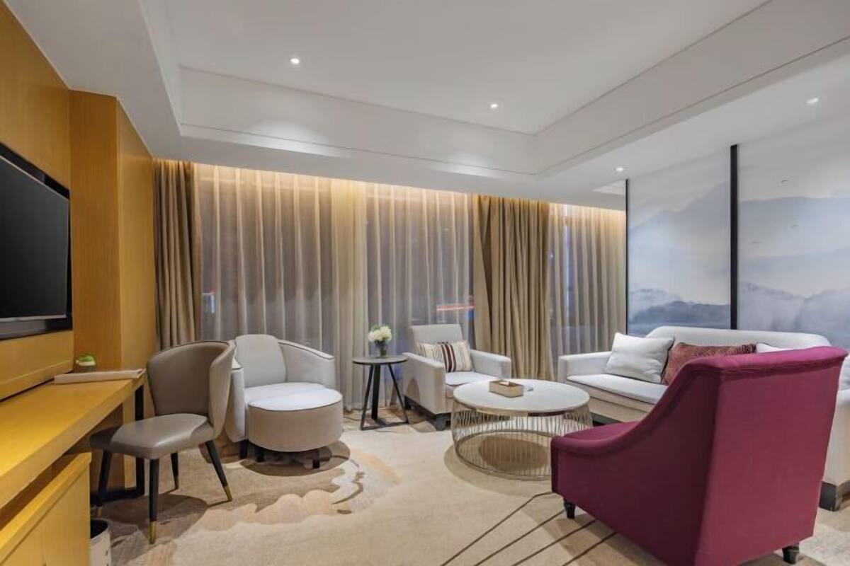 Una habitación de hotel moderna con una televisión de pantalla plana, un escritorio de madera, una mesa de centro redonda, dos sillones, un sofá y un ventanal con cortinas.