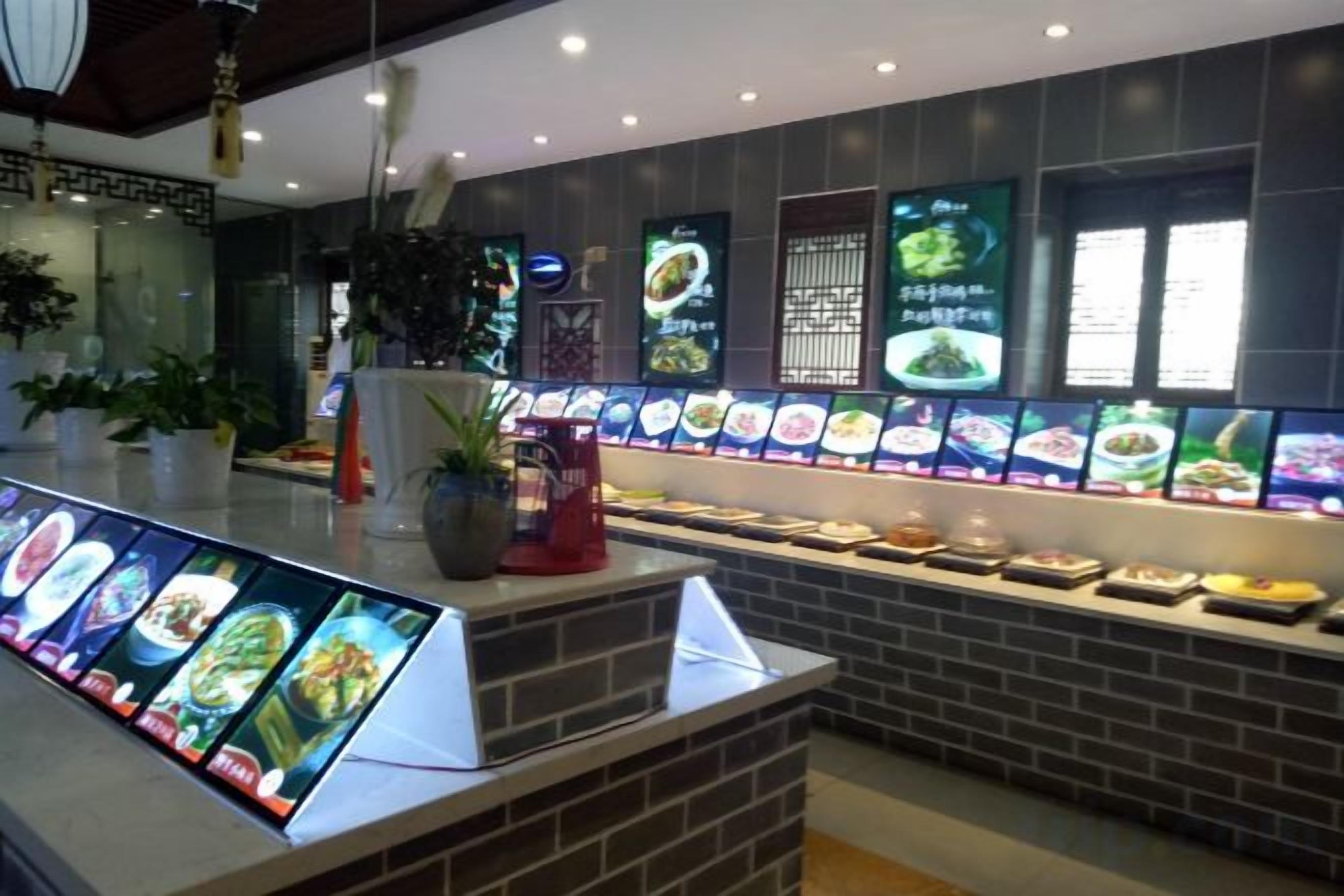 A restaurant interior with a digital menu display, brick wall, and various food items on the counter.