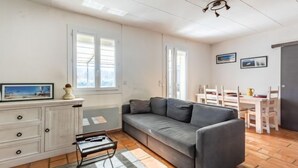 Ein gemütliches Wohnzimmer mit einem grauen Sofa, einem Holzschrank und einem kleinen Tisch. Zwei Fenster sorgen für natürliches Licht und eine Tür führt in einen anderen Raum.