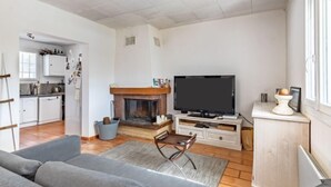 Ein Wohnzimmer mit Kamin, Fernseher und einer Couch. Im Hintergrund ist eine Küche zu sehen.