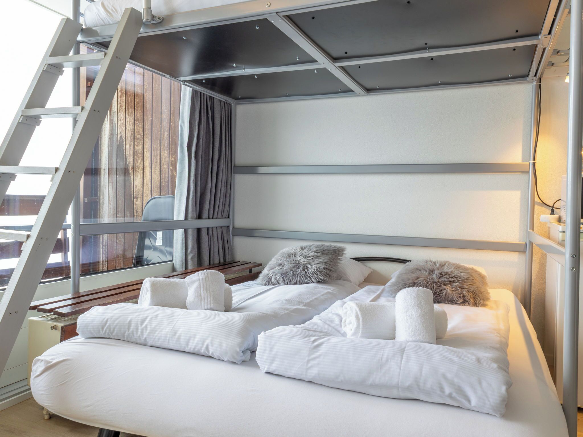 A modern bedroom with a bunk bed, a ladder, a window with curtains, and a bedside table with a lamp.