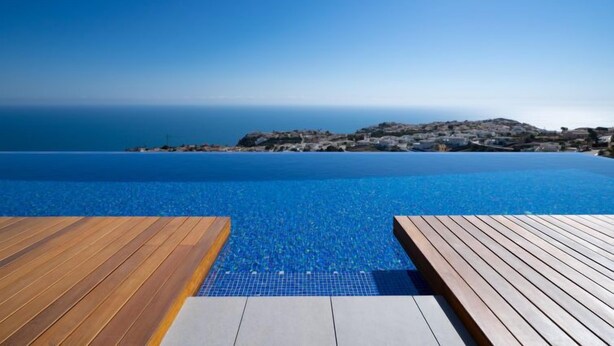 Une piscine d’un bleu éclatant, entourée de terrasses en bois, offrant une vue sur une ville côtière et la mer.