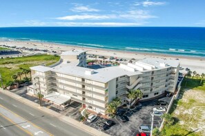 A multi-story hotel with a flat roof, located near a beach with visible parking and a road in the foreground.
