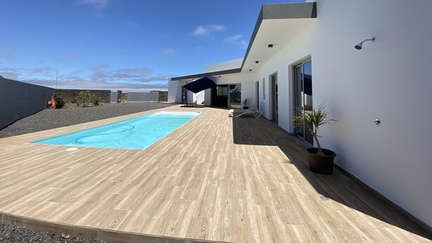 Un área de piscina moderna al aire libre con terraza de madera, zona de estar cubierta y cielo azul despejado.