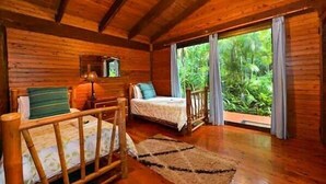 A wooden cabin room with a bed, a small table, and a window overlooking greenery.