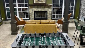 Una habitación con mesa de futbolín, chimenea, sofá y dos ventanas con persianas.