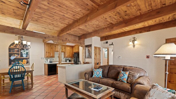 A spacious living area with a wooden ceiling, a leather sofa, a glass coffee table, and a dining area with a blue chair. - Fort Marcy 17 | Walk to Plaza | Fireplace | Kitchen | Hot Tub | Pool | End Unit | Single Level (Santa Fe)