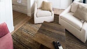 Un salon avec un plafond haut, un téléviseur fixé au mur, une table basse en bois et un fauteuil beige.