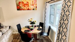 Een eetgedeelte met een ronde tafel, zwarte stoelen en een bloemenvaas. Er is een raam met gordijnen met een patroon en een schilderij van bloemen aan de muur.