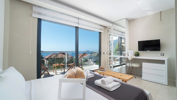 A hotel room with a large balcony, a bed, a desk with a TV, and a view of the sea. - Villa Sole Sidyma – Luxury Villa with Heated Pool, Central Location, Bay Views (Antalya)