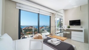 A hotel room with a large balcony, a bed, a desk with a TV, and a view of the sea.