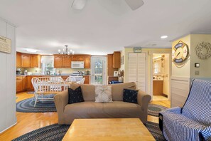 Una sala de estar acogedora con un sofá, una mesa de centro y un sillón a rayas. Se observa una cocina al fondo con armarios de madera y un comedor.