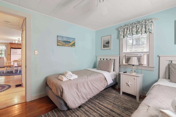 Un dormitorio con cama, mesitas de noche, una ventana con cortinas y una puerta que da a otra habitación.