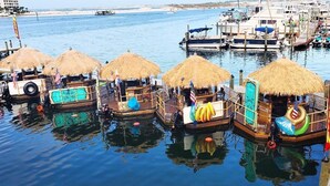 A row of thatched-roof boats docked at a marina with clear blue water and a sandy beach in the background. - 2BR Updated Condo | 2 Pools, Steps to Beach (Miramar Beach)