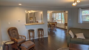 A spacious living room with a wooden floor, a green sofa, a wooden chair, and a dining area with wooden chairs and a table. - 2500 SF Remodeled Cottage on a Quiet Lake in the Woods (Rhinelander)