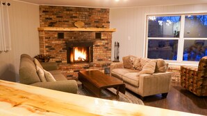 A cozy living room with a brick fireplace, a sofa, a coffee table, and a patterned armchair. - 2500 SF Remodeled Cottage on a Quiet Lake in the Woods (Rhinelander)