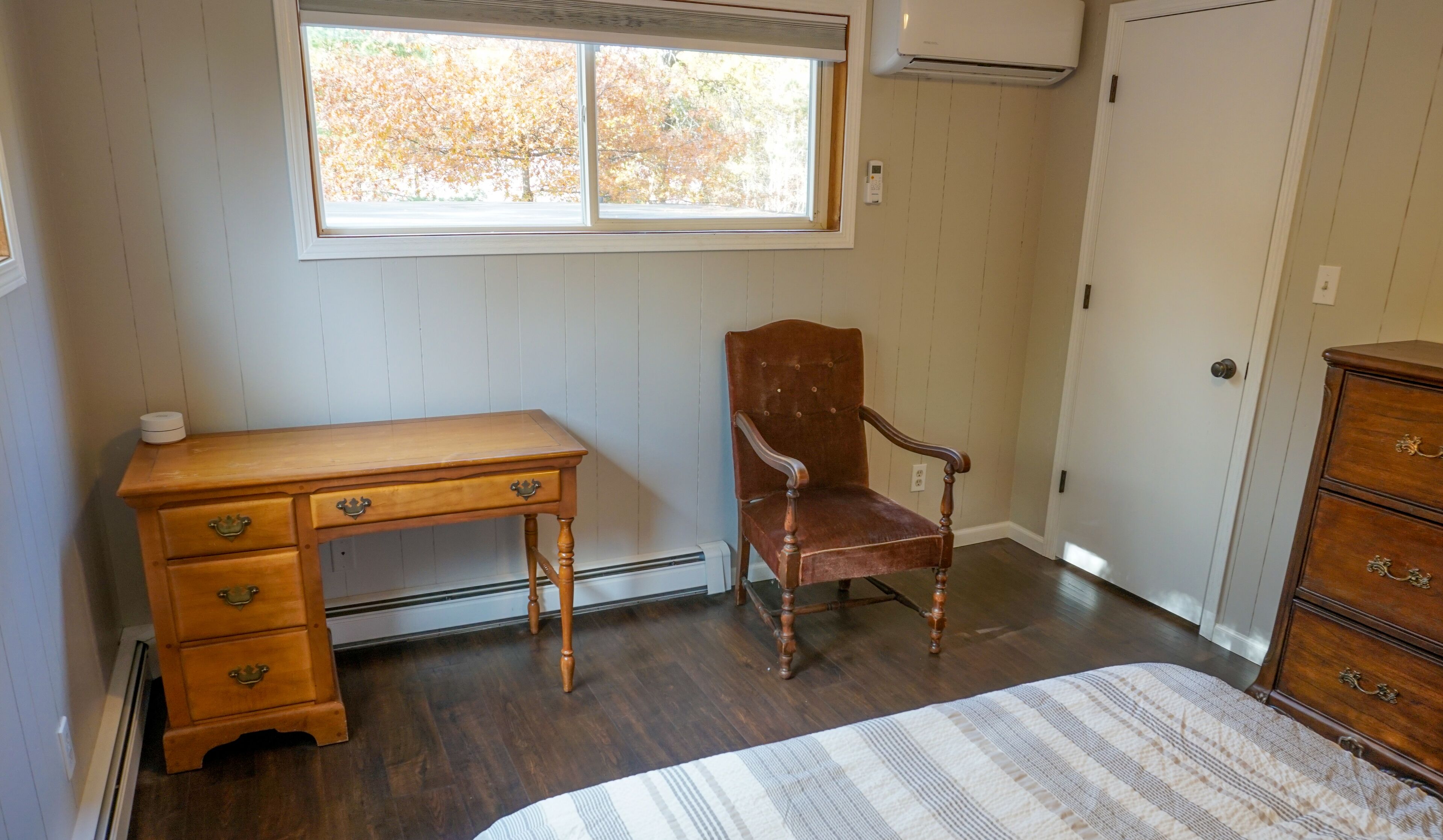 A room with a wooden desk, a chair, a dresser, and a window with a view of trees.