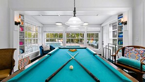 Une table de billard avec des billes et des queues, entourée de chaises et d’étagères remplies de billes.