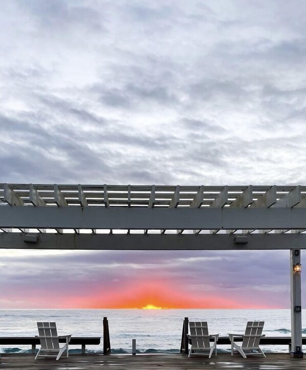 A pier with a metal railing and two empty chairs facing the ocean at sunset.