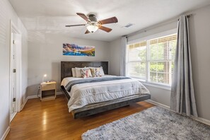 Un dormitorio con una cama, una mesita de noche, un ventilador de techo, una ventana con cortinas y un cuadro en la pared.