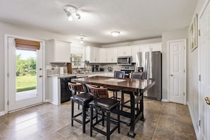 A kitchen with white cabinets, a central island with stools, stainless steel appliances, and a view to the outside.