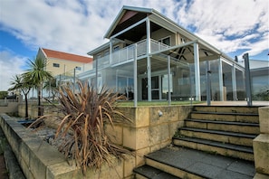 A modern two-story house with a glass balcony, a stone retaining wall, and a palm plant. - “WATERLILY HOUSE” - Luxury Mandurah Canal Holiday Home - Jetty, BBQ, Sleeps 14 (Mandurah)